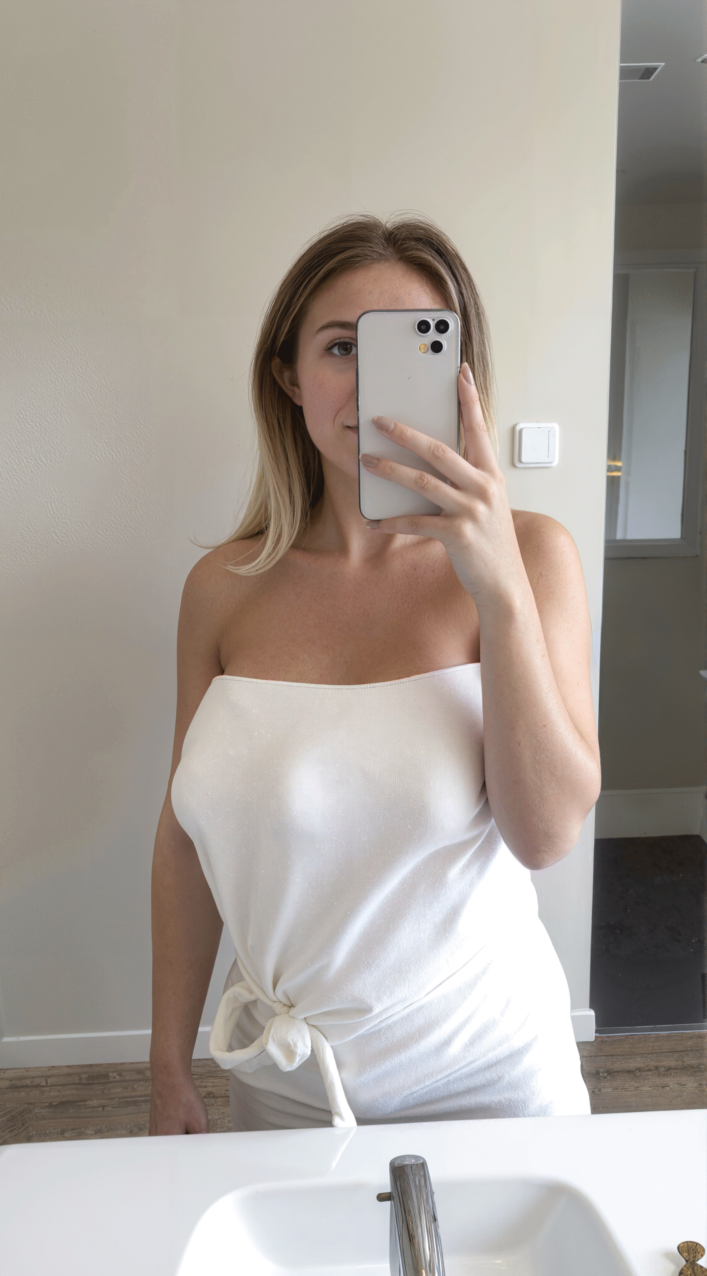 Woman in a white towel dress taking a mirror selfie in a minimalist bathroom